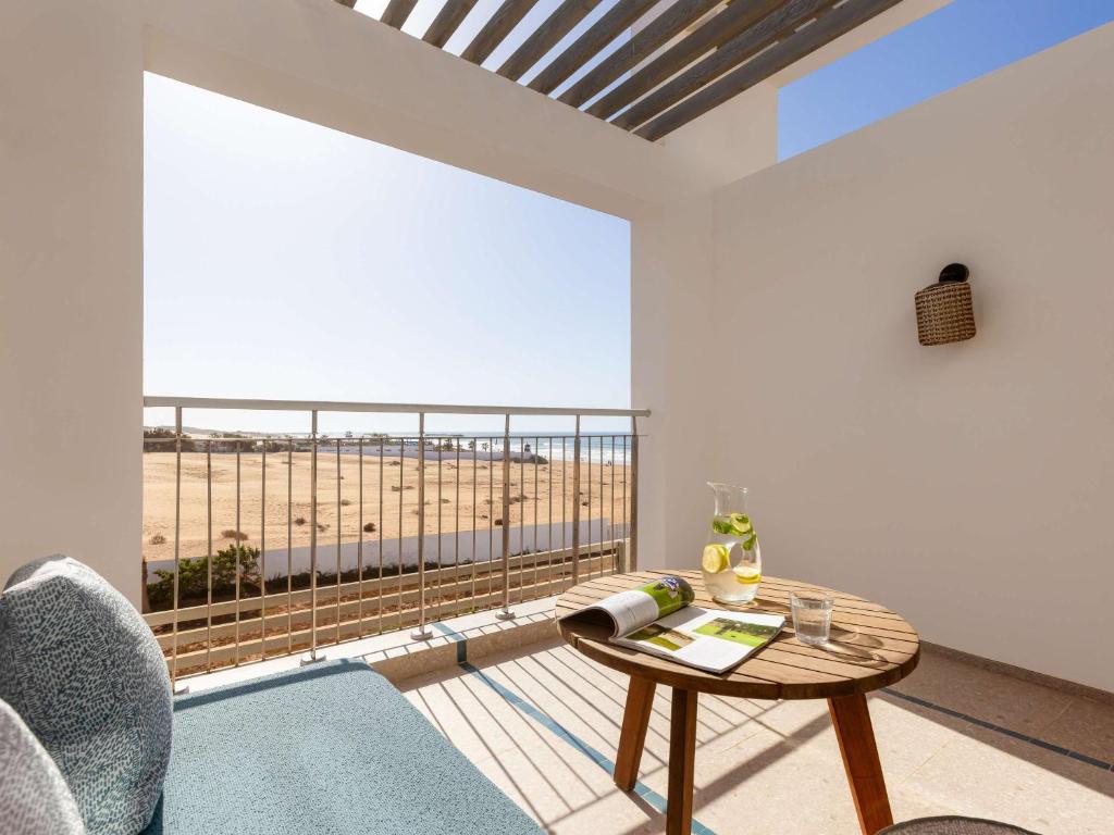 Balkons ar skatu uz pludmali, dekorēts ar modernu mēbelēm un galdu, uz kura atrodas ūdens karafe ar citrona šķēlītēm un grāmata. Rāmā atmosfērā var baudīt dabu un sauli.