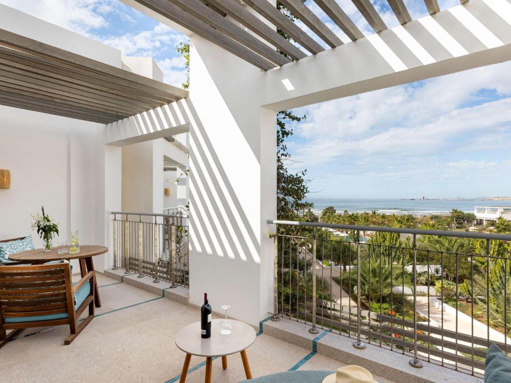 Gaiša terase ar skatu uz jūru, kur atrodas koka mēbeles un dzērienu galds, apzaļumots dārzs un skaidrs debesis.