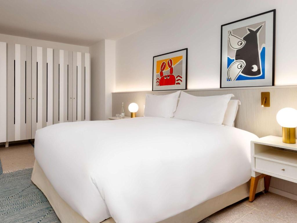 Mūsdienīgs guļamistabas interjers ar plašu gultu, elegantiem mākslas darbiem uz sienām un baltu drēbju skapīti. Gaismas avoti rada siltu atmosfēru.