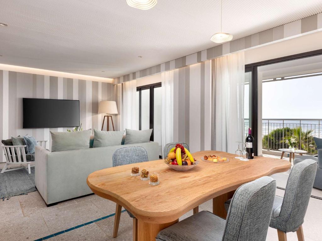 Mūsdienīgs viesistabas un ēdamistabas interjers ar skatu uz jūru, kurā ir koka galda ar augļiem un vīna pudeli, kā arī dīvāns un televizors. Gaismas un neitrālas krāsas rada komfortablu atmosfēru.