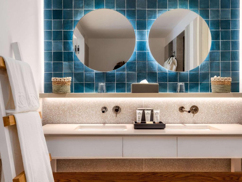 Moderns vannas istabas dizains ar divām apaļām spoguļu atverēm uz tumši zilo flīžu fona, apgaismojuma joslu un elegantu izlietni. Estētika apvieno funkcionalitāti un stilu.