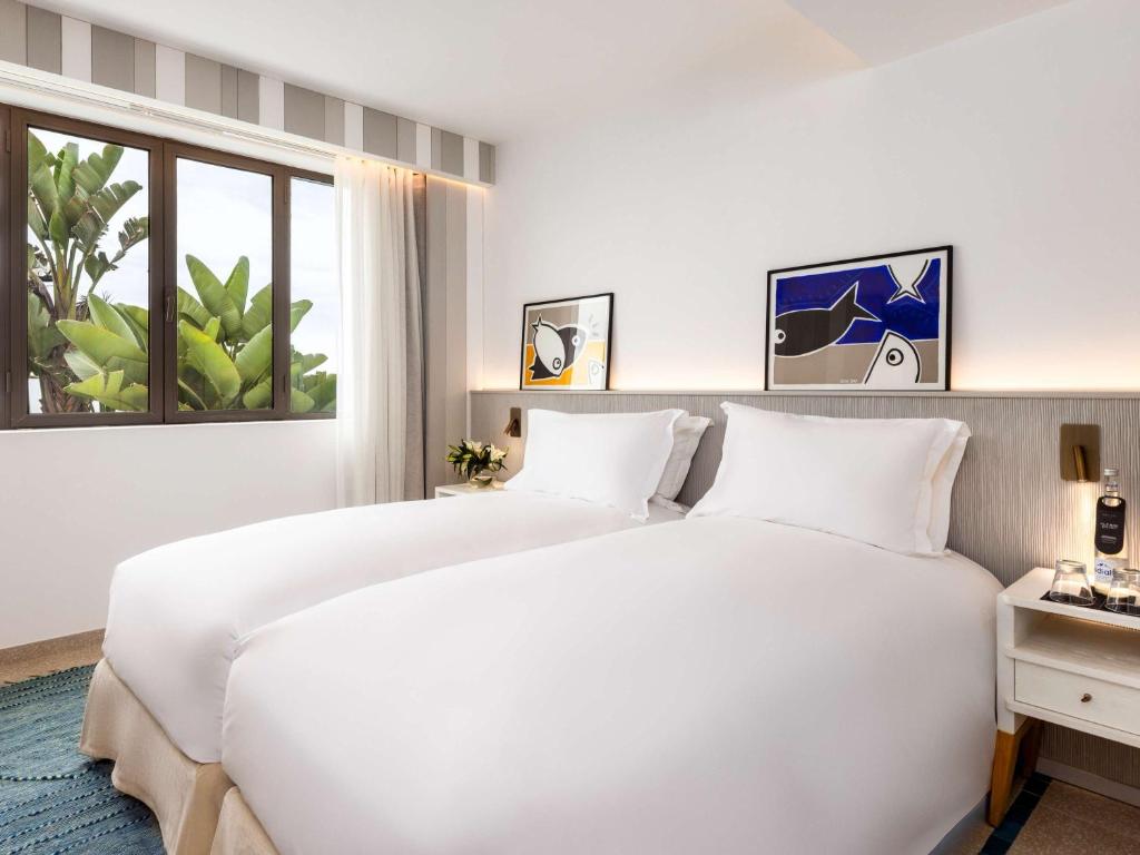 Mūsdienīga viesnīcas numura interjers ar diviem gultas veļas izkārtojumiem, mākslas darbiem uz sienas un plašu logu, kas sniedz skatu uz zaļumu.