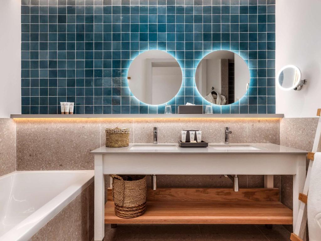 Mūsdienīgs vannas istabas dizains ar tumši zaļiem flīzēm, divām apaļām spoguļiem un elegantu vannas izlietni. Telpā ir siltas gaismas akcenti un dabīgi materiāli.