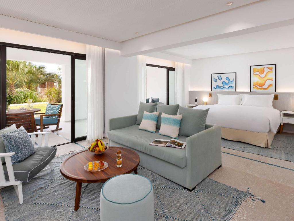 Moderns viesnīcas numurs ar ērtu dīvānu, galdu ar augļiem un skatu uz dārzu. Interjers ir gaišs un elegants, ar mākslas darbiem uz sienām.