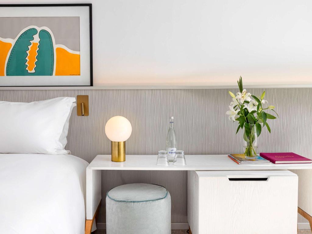 Mūsdienīgs guļamistabas interjers ar baltu gultu, stilīgu galdiņu, lampu un ziedu kompozīciju, kā arī krāsainu mākslas darbu uz sienas.