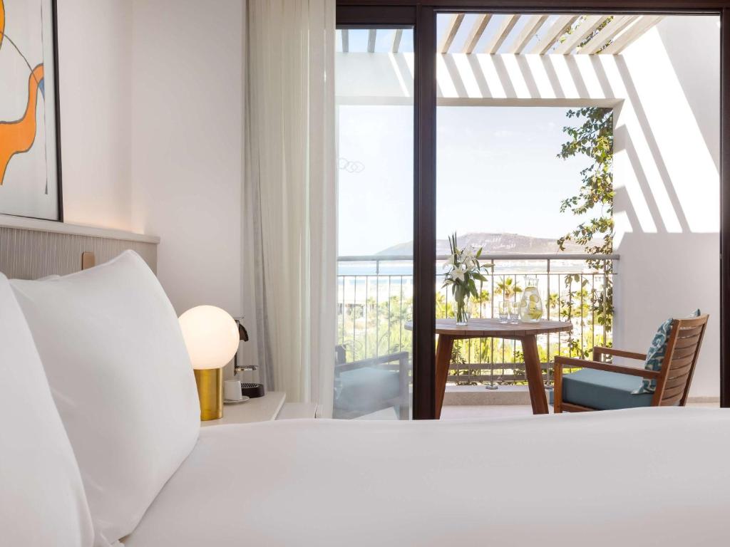 Mūsdienīgs viesnīcas numurs ar skatu uz jūru, kur redzama ērta gulta, moderns apgaismojums un balkons ar galds un krēsls. Ideāla vieta relaksācijai un atpūtai.