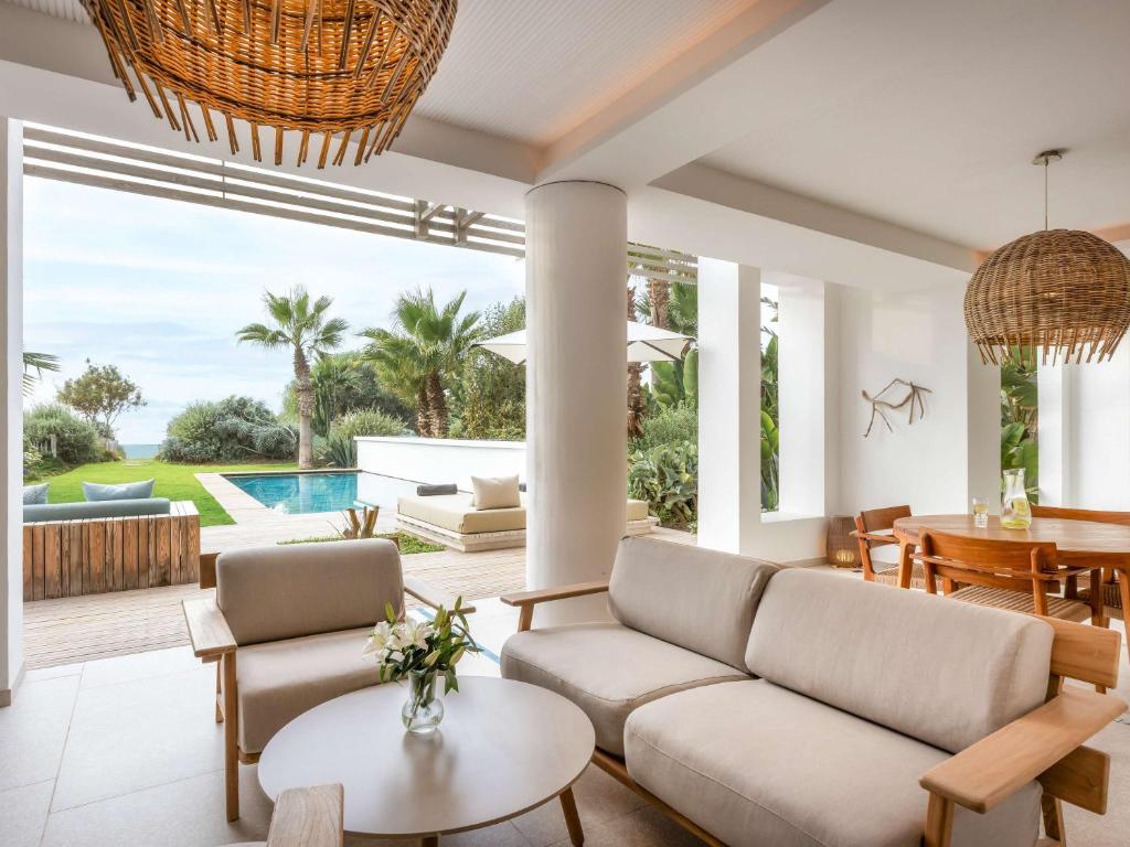 Mūsdienīgs dzīvojamās telpas interjers ar skatu uz dārzu un baseinu, kurā ir ērti dīvāni, koka galdi un stilīgas lampas. Terase ir apzaļumota ar palmām un citām augiem, radot relaksējošu atmosfēru.