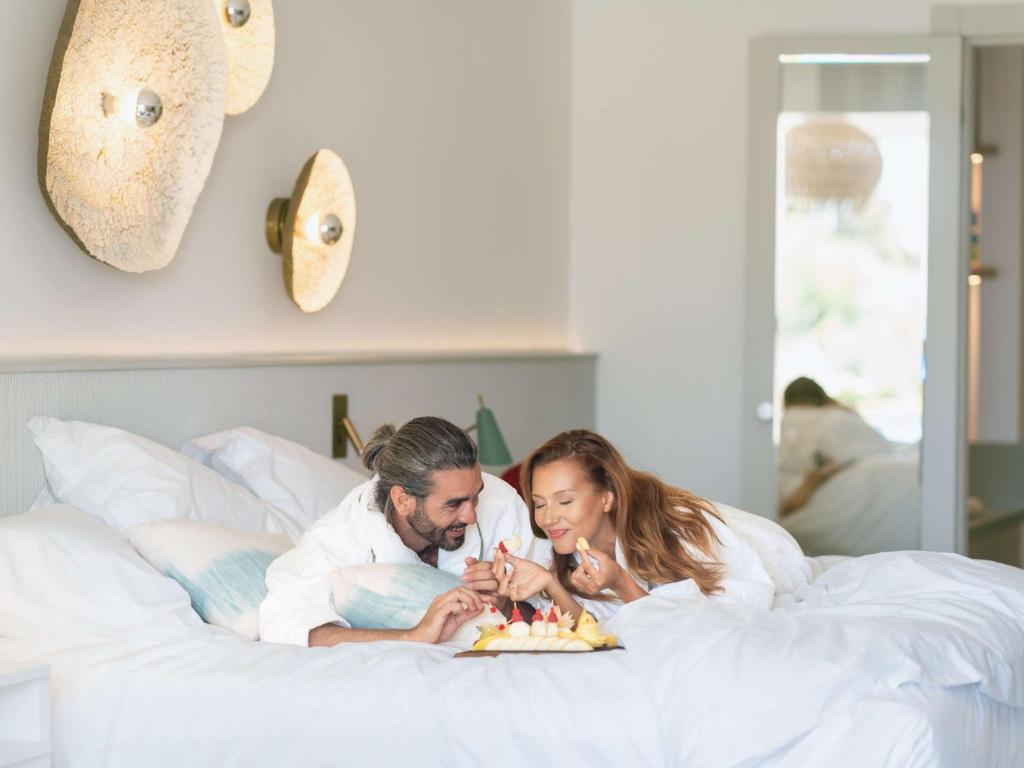 Pāris guļ gultā luksusa viesnīcā, baudot augļus un saldumus. Svaiga un relaksējoša atmosfēra, kas iedvesmo romantiskai atpūtai.