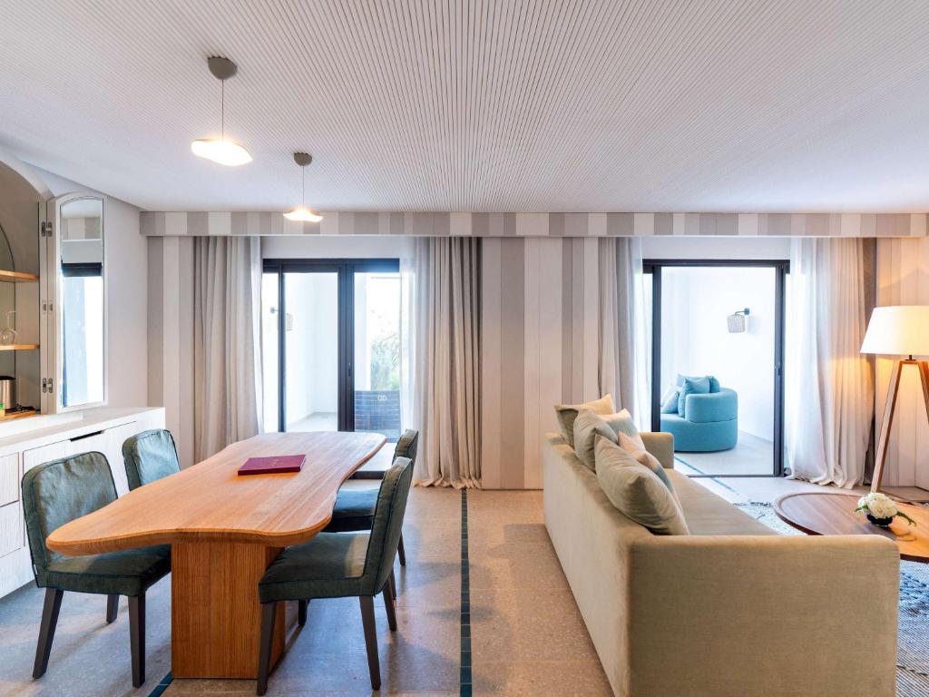 Mūsdienīga viesistaba ar ēdamgaldu un komfortablu dīvānu, gaišām krāsām un lieliem logiem, kas nodrošina dabisko apgaismojumu. Interjers ir elegants un gaumīgs, radot mājīgu atmosfēru.