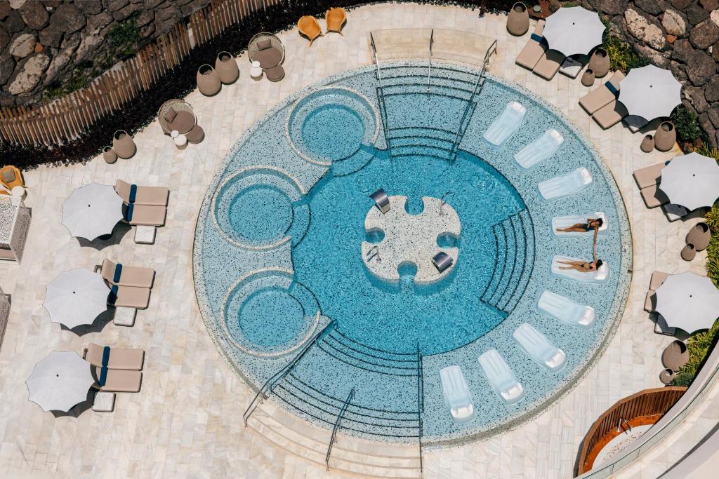 Lieliska atpūtas zona ar apaļu baseinu, kas ieskauts sauļošanās krēsliem un lietussargiem. Baseina centrā redzamas atpūtas gultas, nodrošinot relaksējošu atmosfēru.