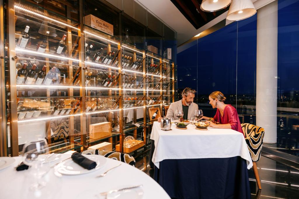Romantiska vakariņas elegantā restorānā ar skatienu uz pilsētu, kur pāris bauda vakariņas pie galda, kas ierīkots blakus vīna glabātuvēm.