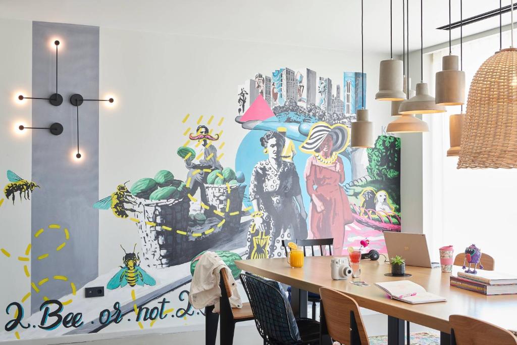 Mūsdienīga interjera telpa ar krāsainu sienu gleznojumu, kas attēlo stilizētus tēlus un dabas elementus. Uz galda ir grāmatas, datori un dzērieni, radot radošu un relaksējošu atmosfēru.