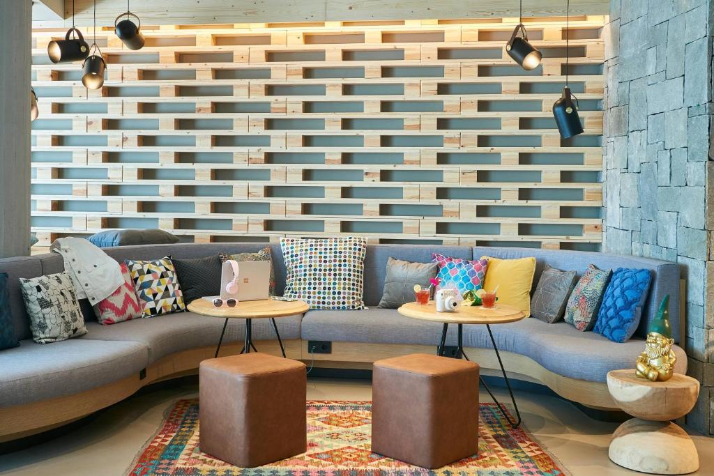 Mūsdienīgs atpūtas stūrītis ar pelēku dīvānu, krāsainām spilveniem un koka sienu, kas piešķir siltu atmosfēru. Divi galdiņi ar dzērieniem un dekoratīviem elementiem, radot patīkamu un viesmīlīgu vidi.