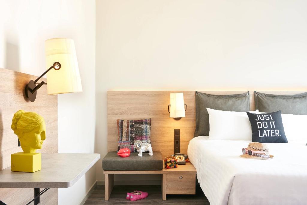 Mūsdienīgs viesnīcas numurs ar ērtu gultu, stilīgu apgaismojumu un dekoratīviem elementiem, piemēram, dzeltenu skulptūru un spilvenu ar uzrakstu. Numurā ir arī mīksta dīvāna zona un neliels galdiņš.