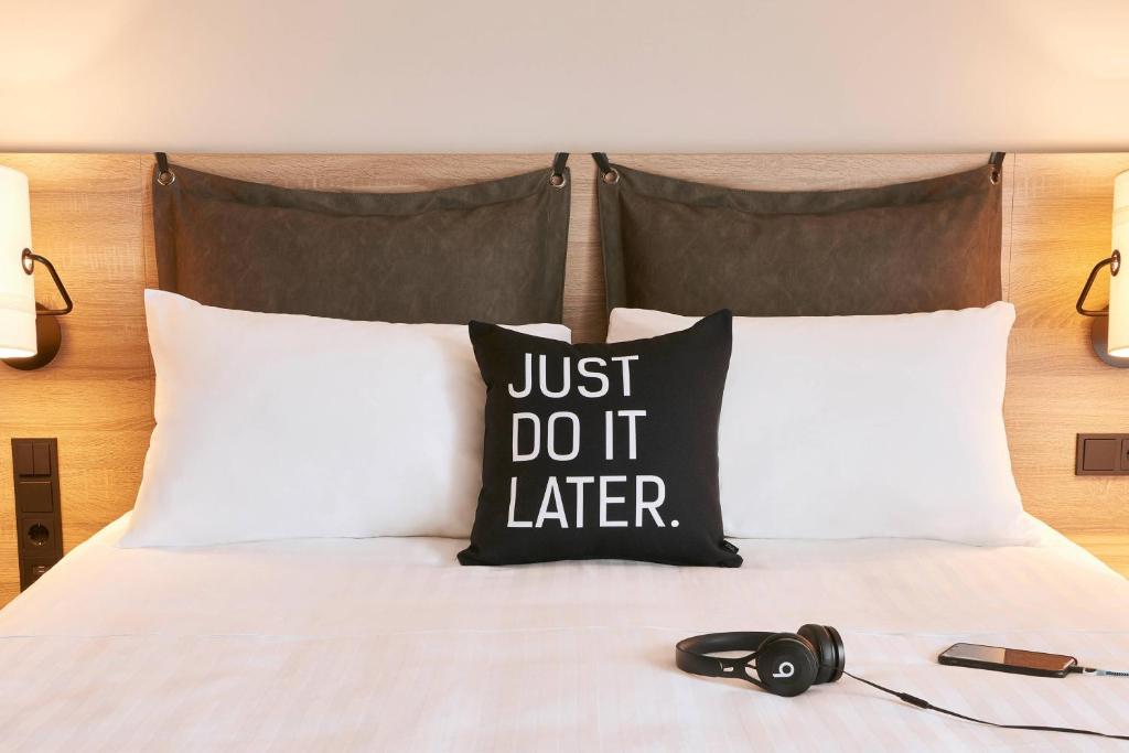 Gultas spilvens ar uzrakstu "JUST DO IT LATER" un baltiem spilveniem, blakus atrodas austiņas un telefons. Interjers ir moderns un mājīgs.