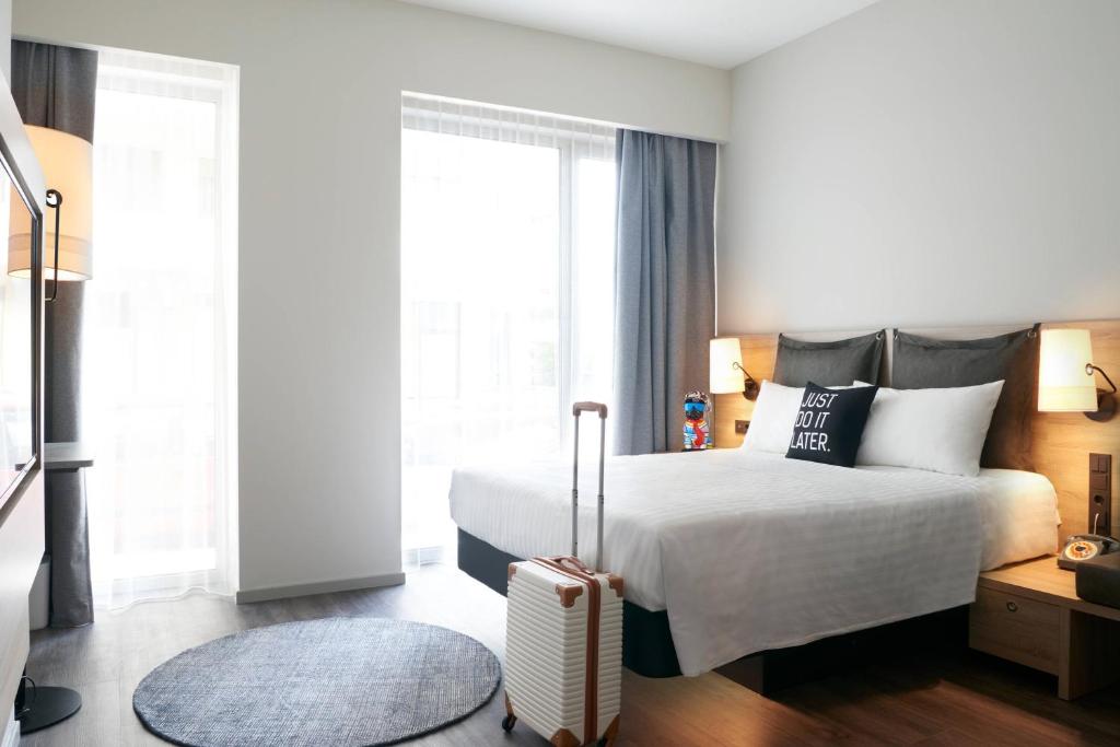 Mūsdienīga viesnīcas istaba ar divvietīgu gultu, mīkstiem spilveniem un bagāžu. Gaismas un logi rada gaišu un ērtu atmosfēru.