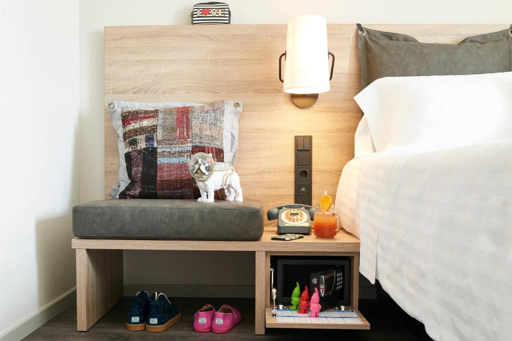 Mūsdienīga guļamistabas interjera kompozīcija ar stilīgu gultas galvgali, mīksto sēdi un dažādiem aksesuāriem, tostarp apģērbu un dekoratīvu suņu figūru.