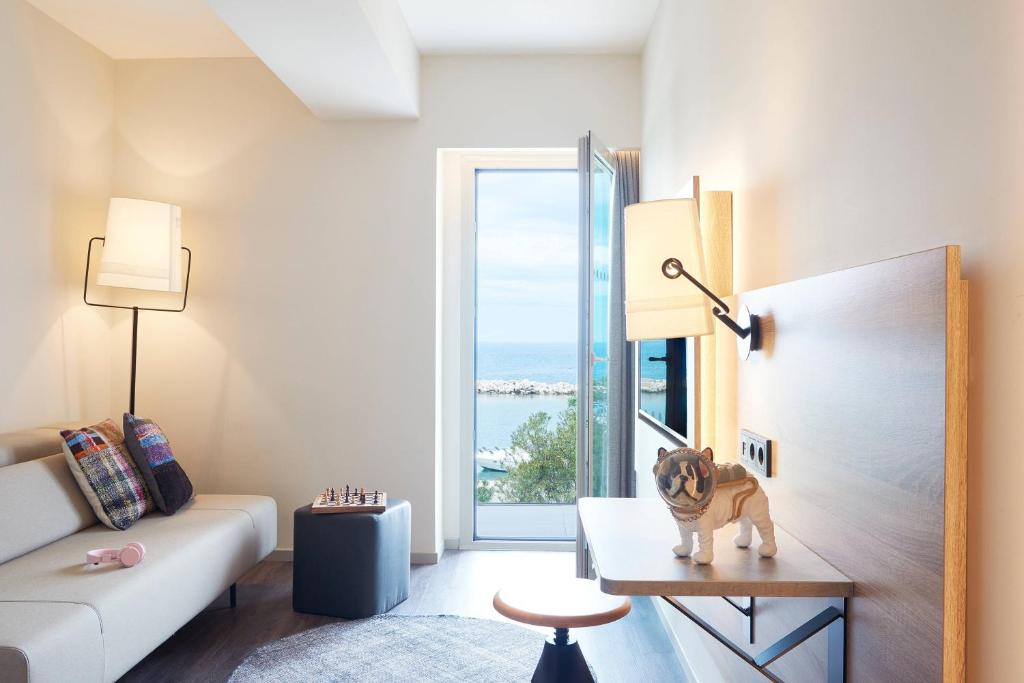 Mūsdienīgs viesnīcas numurs ar skatu uz jūru, iekārtots ar ērtu dīvānu, stilīgu galdu un dekoratīvu suņu figūru. Interjers ir gaišs un elegants, radot patīkamu atmosfēru.