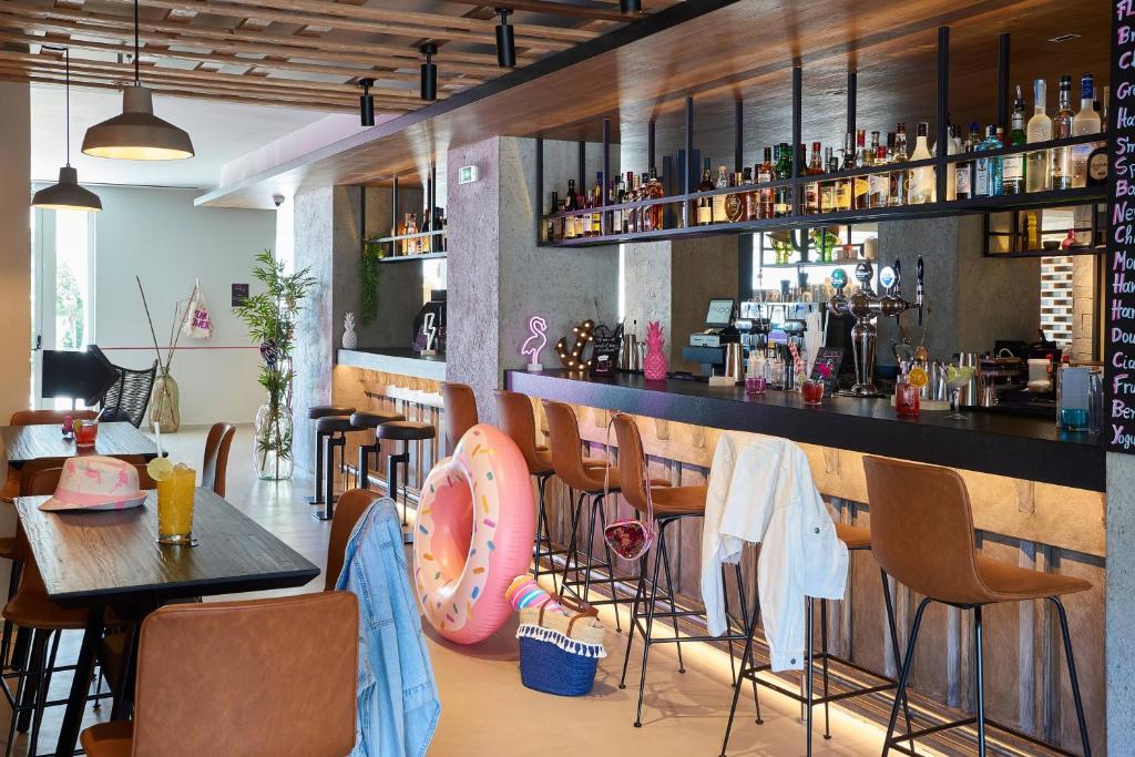 Mūsdienīgs bārs ar elegantu interjeru, koka grīdu un plašu dzērienu izvēli. Uz galda atrodas kokteiļi un vasaras aksesuāri, kas piešķir jautru atmosfēru.