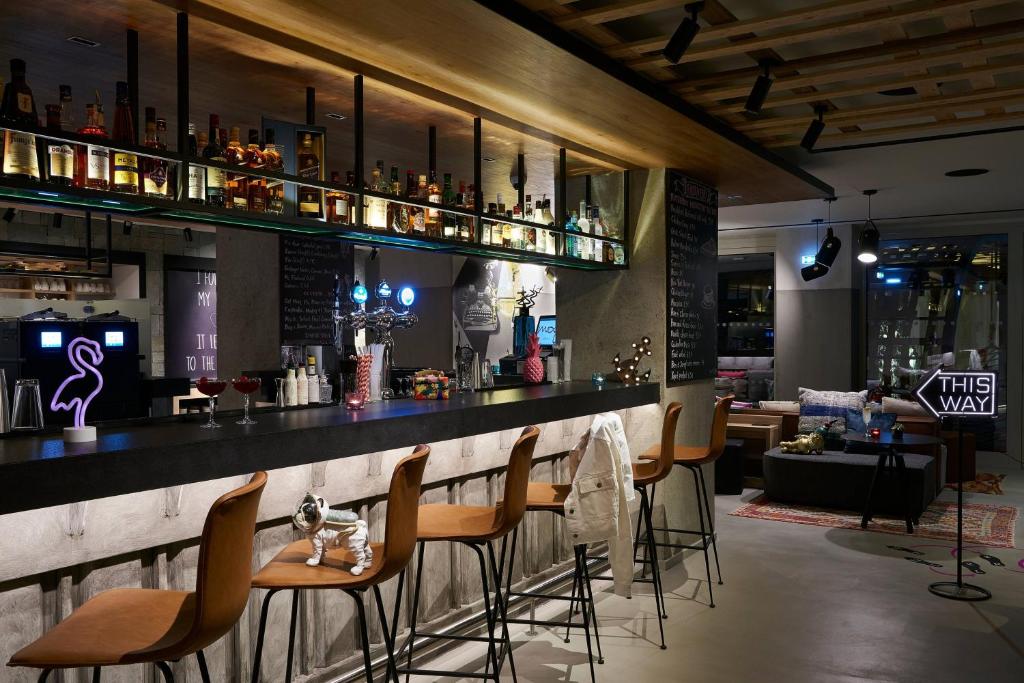 Mūsdienīgs bārs ar elegantu interjeru, kas ietver stilīgas krēslus un radošas dekora elementus. Bāra plauktā izvietoti dažādi dzērieni un gaismas, radot patīkamu atmosfēru.