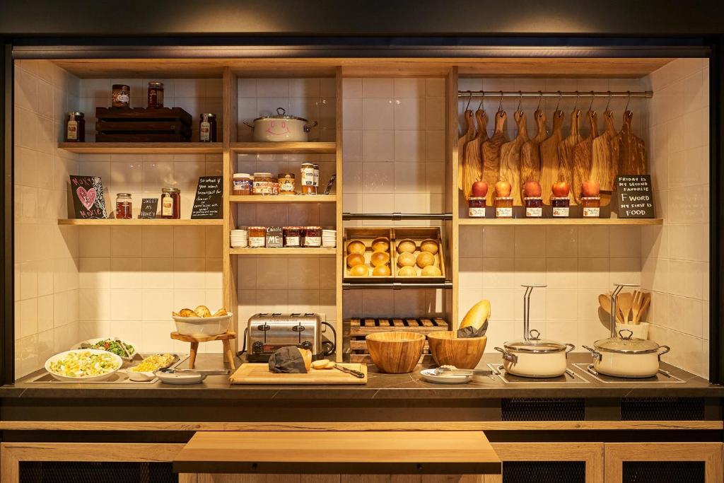 Mūsdienīga virtuve ar koka plauktiem, kur izvietoti dažādi pārtikas produkti, ievārījumi un maizes. Uz galda redzami salāti, trauki un virtuves piederumi, radot mājīgu atmosfēru.