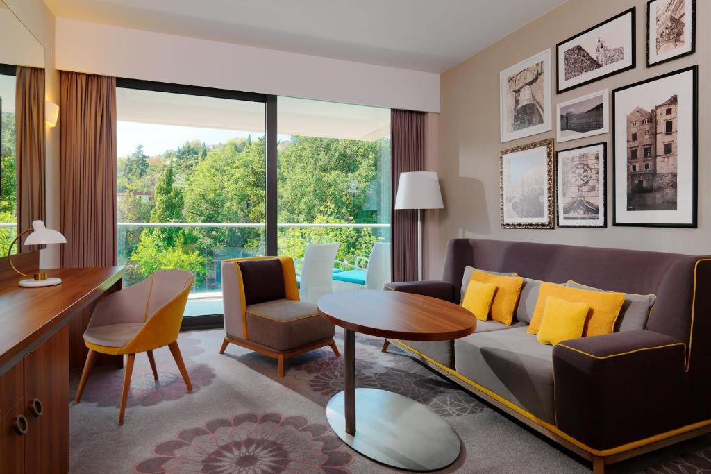 Mūsdienīgs viesnīcas numurs ar komfortablu dīvānu, koka galdu un lieliem logiem, caur kuriem skatās uz zaļojošu ainavu. Sienas rotā dažādas fotogrāfijas un mākslas darbi.