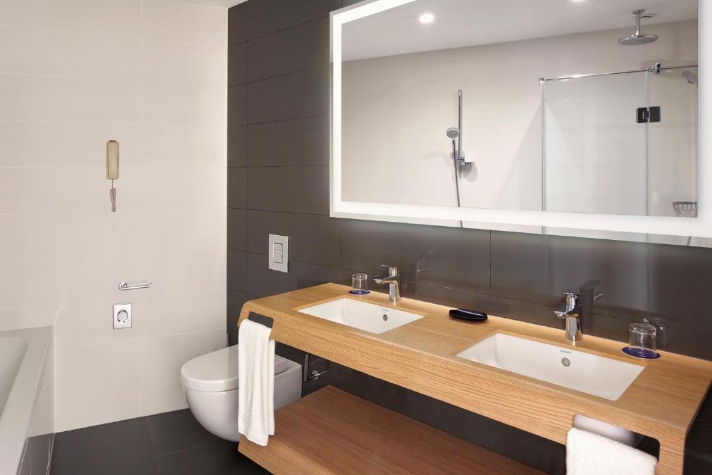 Mūsdienīgs vannas istabas interjers ar divām izlietnēm, koka plauktu un stikla dušu. Iekārtots ar elegantiem santehnikas izstrādājumiem un neitrālām krāsām, radot relaksējošu atmosfēru.