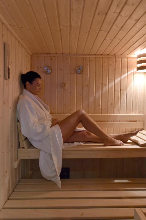 Sieviete baltā halātā relaksējas koka saunā, sēžot uz koka plaukta. Apgaismojums rada mierīgu atmosfēru, uzsverot saunas koka apdari.