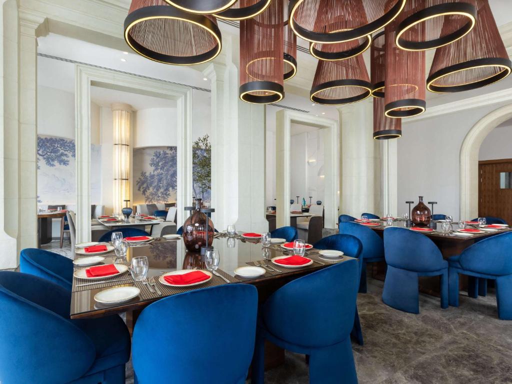 Elegants restorāna interjers ar zilu krēslu un modernu apgaismojumu, kas radīts mājīgas atmosfēras veidošanai. Uz galdiem ir sarkani trauki un glāzes, kas piešķir dzīvīgumu.