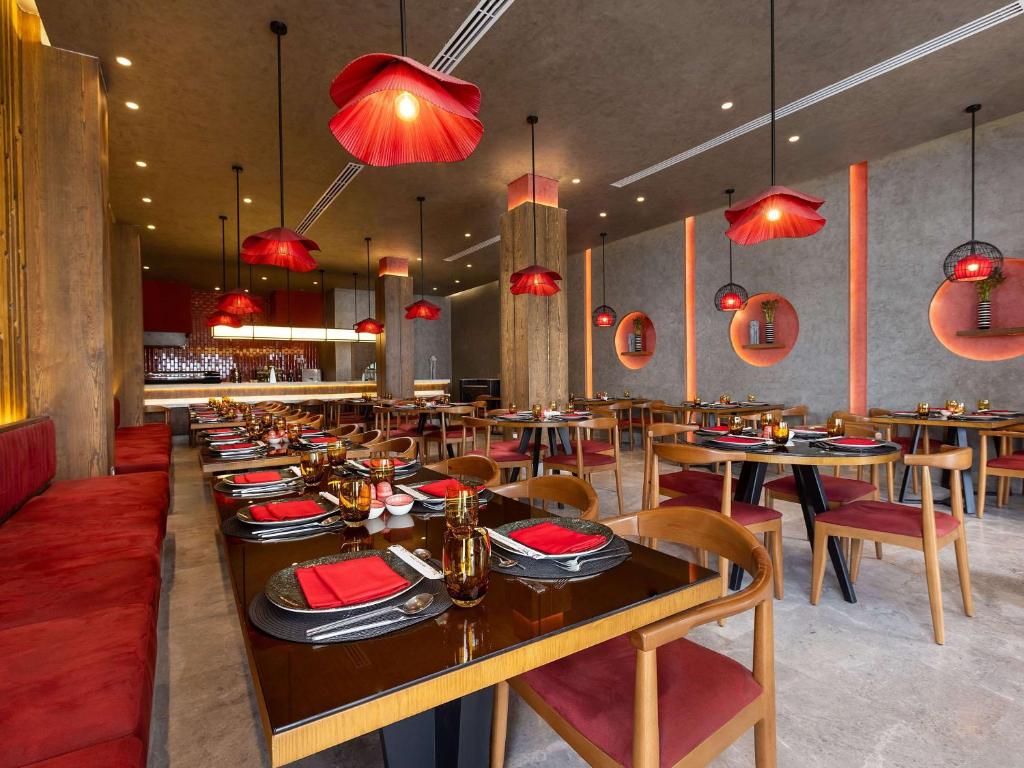 Mūsdienīgs restorāns ar elegantiem sarkaniem apgaismojuma elementiem un stilīgu interjeru. Galdi ir sakārtoti ar melniem šķīvjiem un sarkaniem galdautiem, radot siltu un viesmīlīgu atmosfēru.