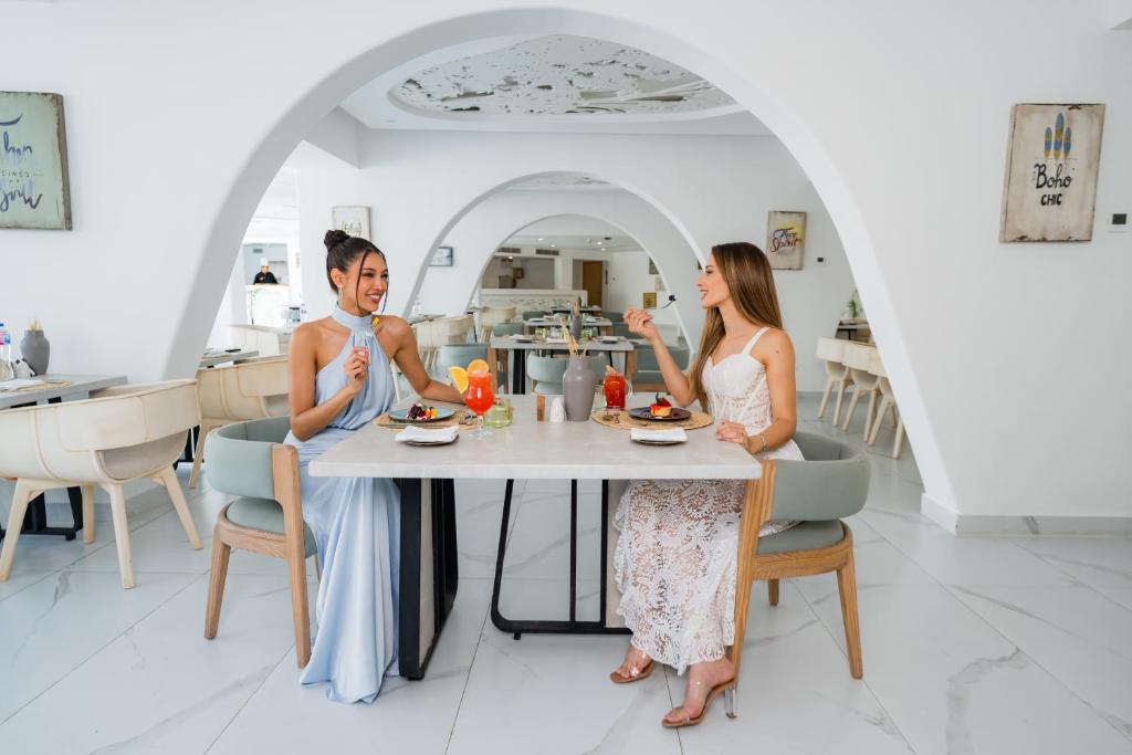Divas sievietes sēž modernā restorānā pie galda, baudot ēdienu un dzērienus, ar elegantu interjeru un gaišām krāsām.