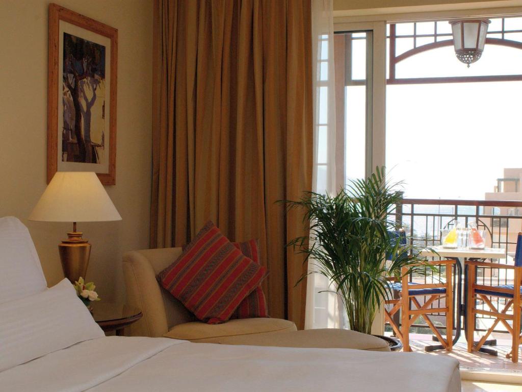 Mūsdienīga viesnīcas numura interjers ar gultu, apgaismojumu un dīvānu, kas piedāvā piekļuvi balkonam ar skatu uz ārpusi. Skatā redzama zaļa auga un galdiņš ar krēsliem.