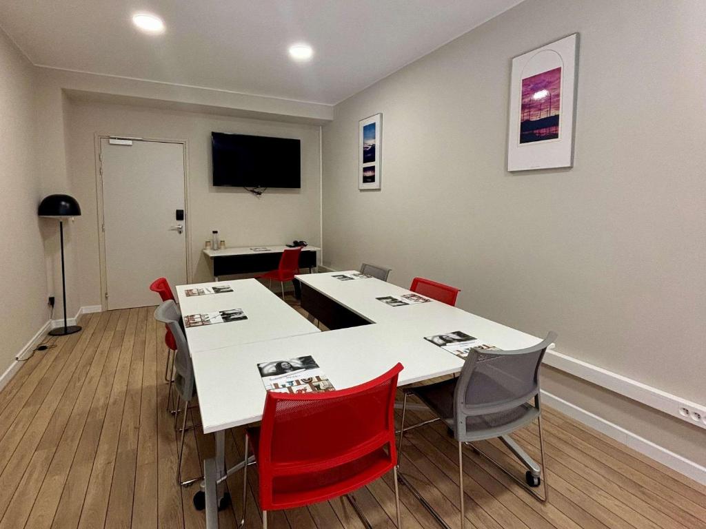 Mūsdienīga konferenču telpa ar baltu galdu un krāsainām krēslām, televizoru un dekoratīvām gleznām uz sienas. Telpa ir gaiša un funkcionāla, piemērota sanāksmēm un izglītojošām aktivitātēm.