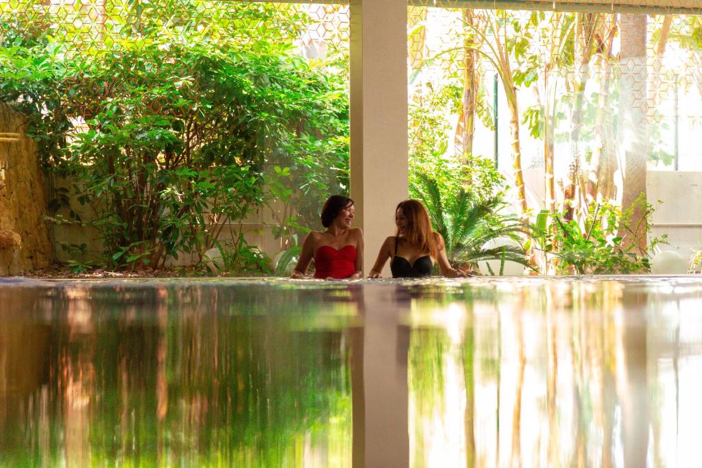 Divas sievietes atpūšas baseinā, apņemti ar zaļām augiem un saules gaismu, radot relaksējošu atmosfēru.