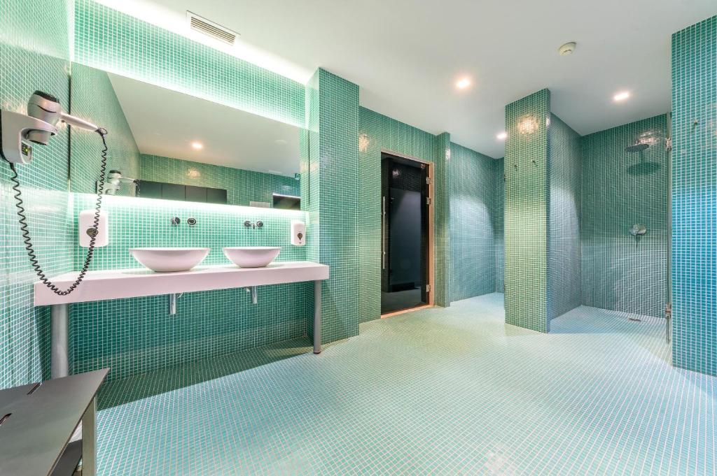 Mūsdienīgs un stilīgs vannas istabas dizains ar zaļām mozaīkas flīzēm, diviem izlietņu traukiem un modernām dušām. Telpa ir gaiša un plaša, radot relaksējošu atmosfēru.
