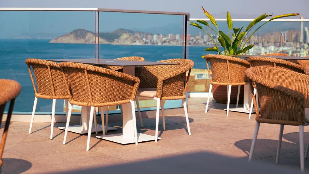 Terases ēdamzona ar vītņotām krēsliem un skatu uz jūru. Aiz krēsliem redzama zaļa auga kompozīcija un pilsētas ainava.