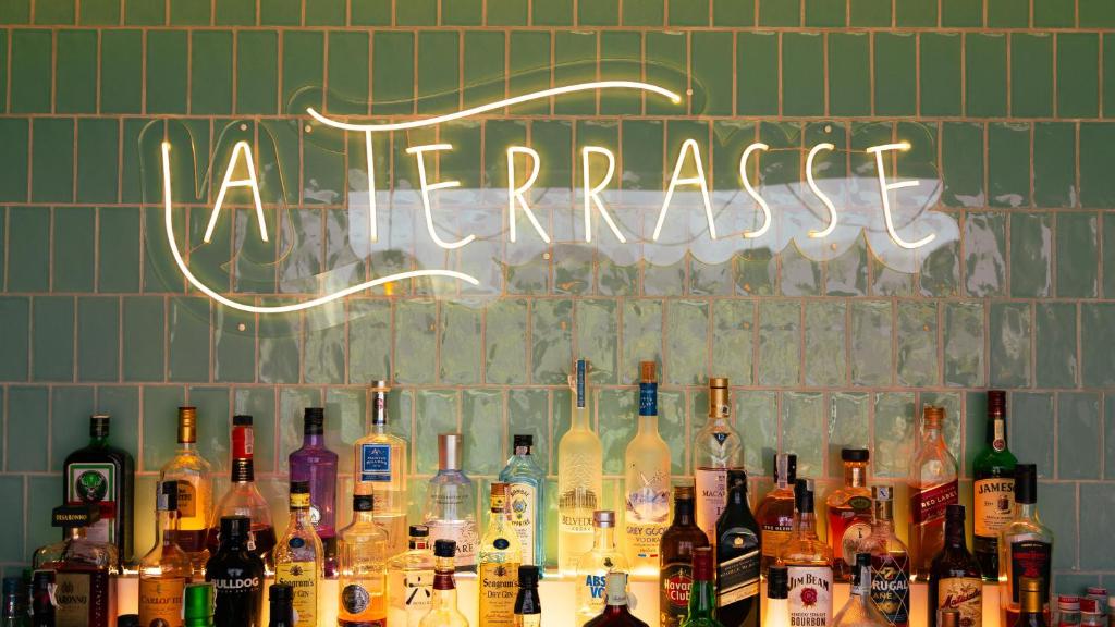 Bāra "La Terrasse" neon gaisma uz zaļām flīzēm, ar dažādām alkohola pudelēm.