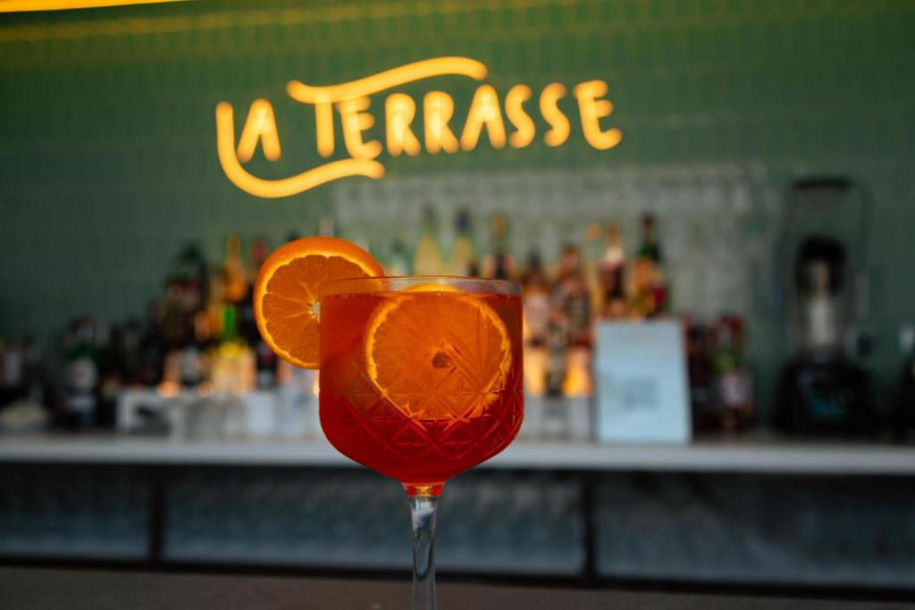 Skaista kokteiļa glāze ar apelsīnu šķēlēm, fona bārā redzami dzērieni un "La Terrasse" nosaukums izceltā apgaismojumā.