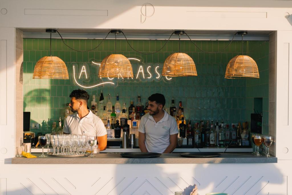 Bāra skats ar diviem bārmeņiem, kas strādā pie modernā bāra ar zaļiem flīzēm un rotājošām lampām. Aiz bāra redzami dažādi dzērieni un glāzes, radot dzīvīgu atmosfēru.