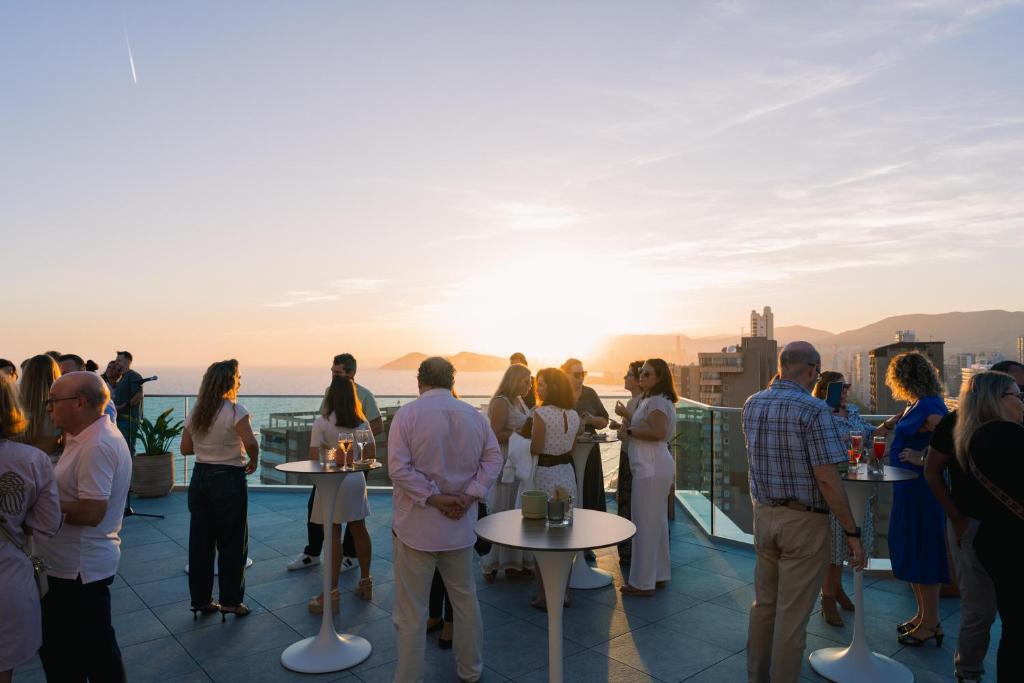 Saulrieta ballīte uz jumta ar cilvēkiem, kas bauda dzērienus un sarunas, fona jūra un pilsētas ainava.