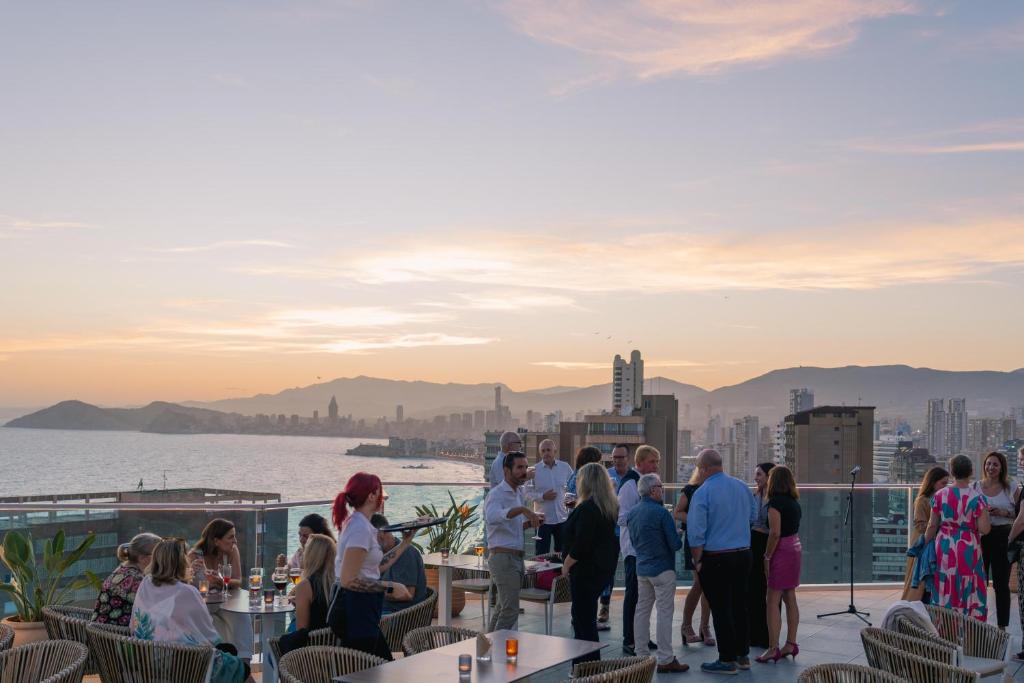 Skaista saulrieta skats no jumta terases, kur cilvēki socializējas un izbauda dzērienus. Fons veido pilsētas panorāma ar kalniem un jūru.