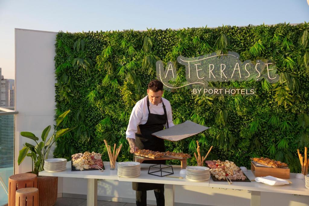 Svaigā gaisā, šefpavārs sagatavo gardumus uz terases, ko rotā zaļa auga siena un stilīga "La Terrasse by Port Hotels" zīme.