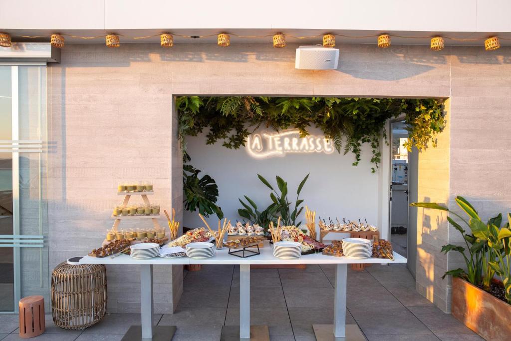 Dekorēts ēdināšanas galds ar dažādām uzkodām un dzērieniem, apgaismojums un zaļumi radījuši patīkamu atmosfēru terasē. Ievērojams interjera elements ir gaismas uzraksts "Terrasse".