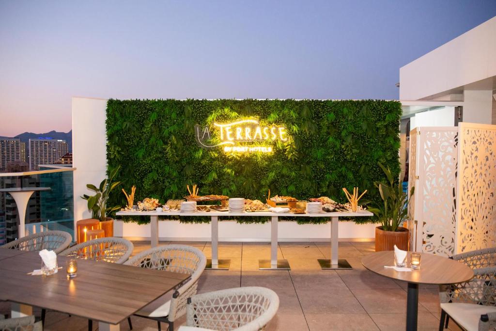 Elegantā terase ar zaļām sienām un apgaismojumu, kurā redzams bufete ar ēdieniem, ideāla romantiskai vai draugu vakariņai. Mājas atmosfēra un pilsētas skats.