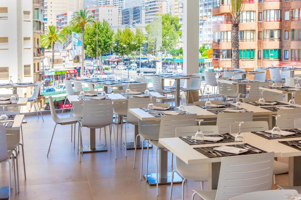 Mūsdienīgs restorāns ar stilīgu interjeru un skatu uz pilsētas ielu, kurā redzami balti galdi un krēsli. Gaismas pieplūdums no logiem rada patīkamu atmosfēru.