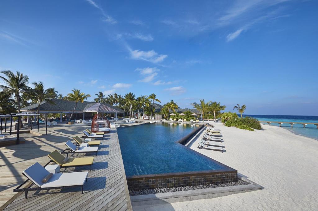 Tropu pludmales kūrorts ar baseinu, sauļošanās krēsliem un palmām, kas apņem relaksējošu vidi. Skats uz zilo jūru un debesi, radot mierīgu atmosfēru.