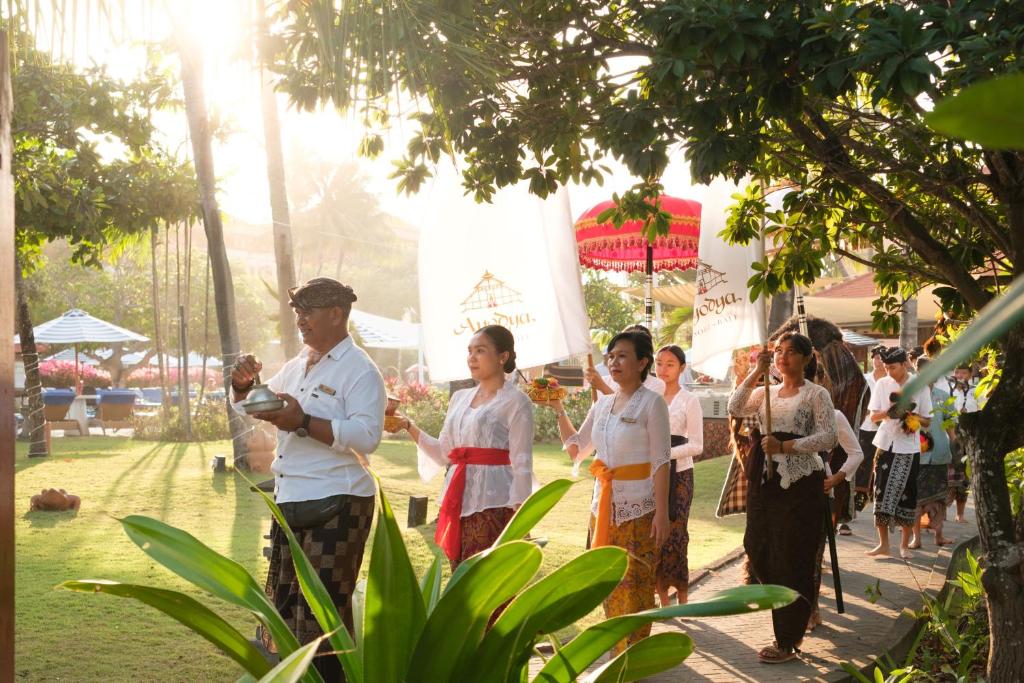 Bali svinību gājiens ar tradicionāliem tērpiem un rotājumiem, saulainā vidē. Gājienā piedalās cilvēki, kas nes karogus un ziedus, radot svinīgu atmosfēru.