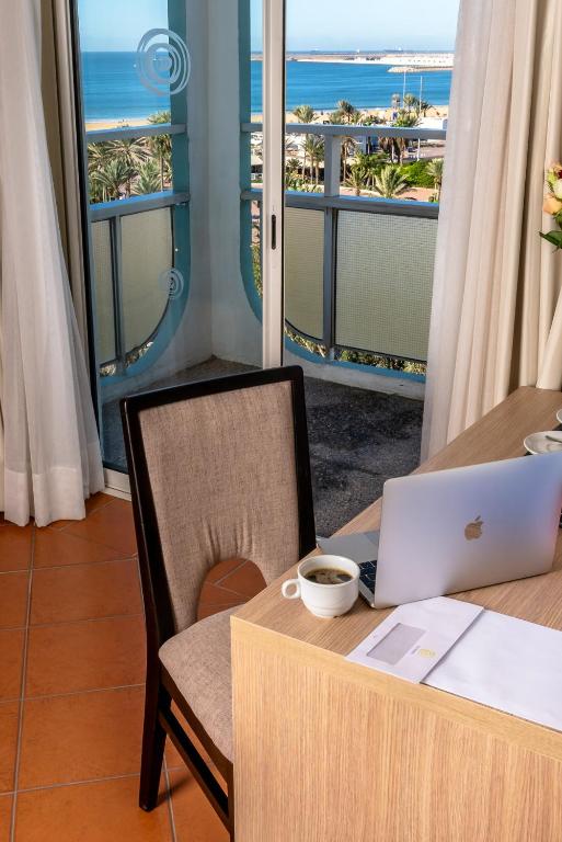 Balcony view from a hotel room with a workspace featuring a laptop, coffee cup, and documents, overlooking the beach and palm trees.