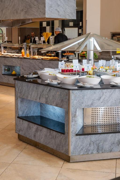 Moderns bufetes galds ar dažādiem ēdieniem un salātiem, kas izvietoti baltos traukos. Restorāna fons rāda darbiniekus, kas gatavo ēdienus.