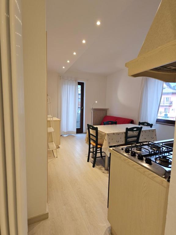 Gaiša virtuve ar ēdamgaldu, krēsliem un sarkano dīvānu, kas redzama caur logiem ar aizkariem. Telpa ir moderna un ērta, ar koka grīdu un gaismas elementiem.